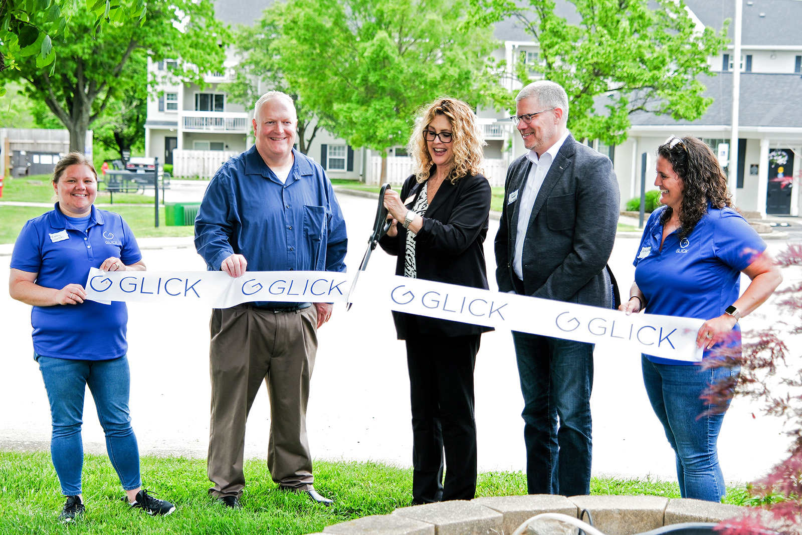 A group of smiling people holding a banner attend a ground breaking ceremony while a woman with large scissors cuts the banner.