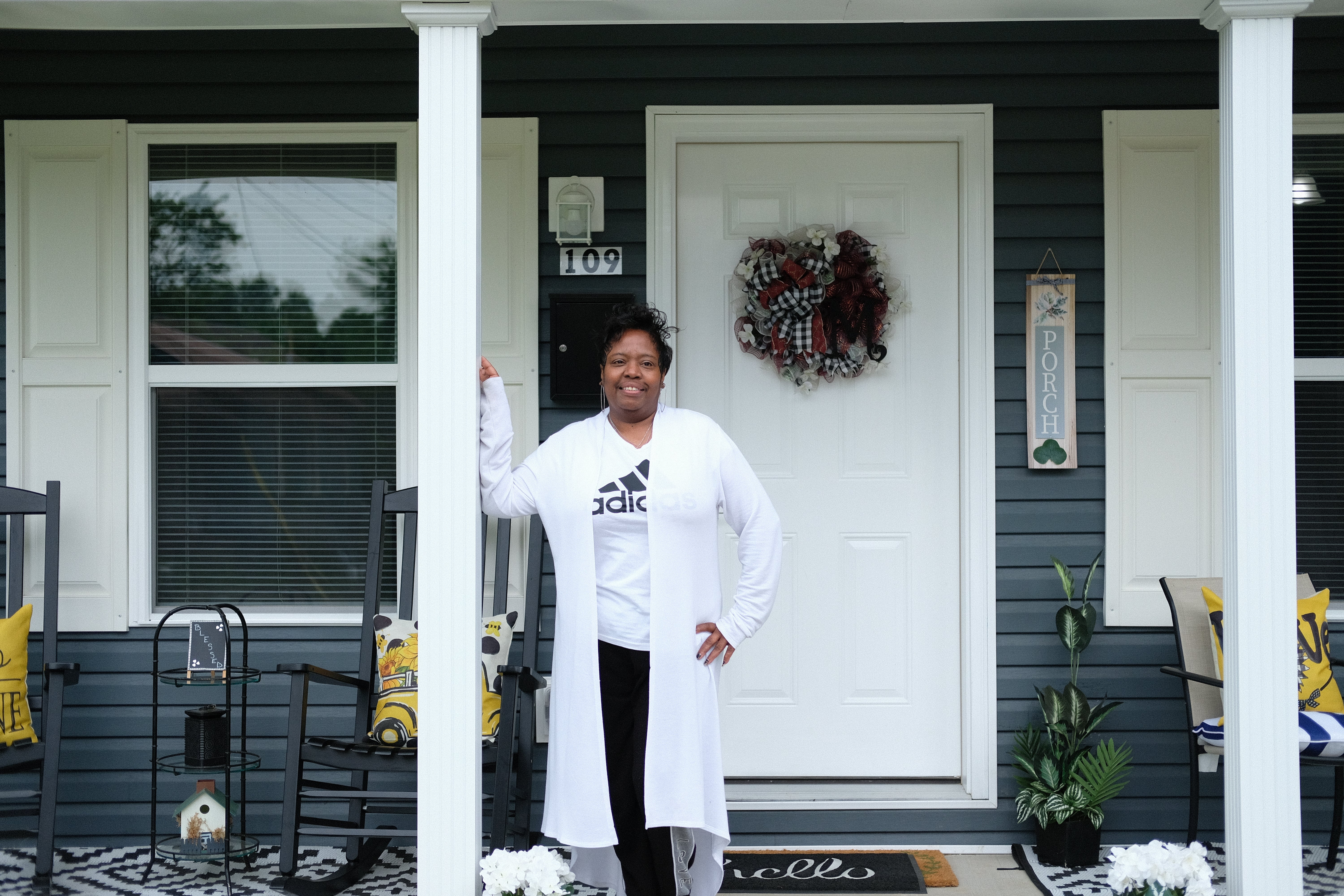 A woman stands on the front porch of a house, leaning against a white pillar, with outdoor seating and a decorative wreath on the door behind her.