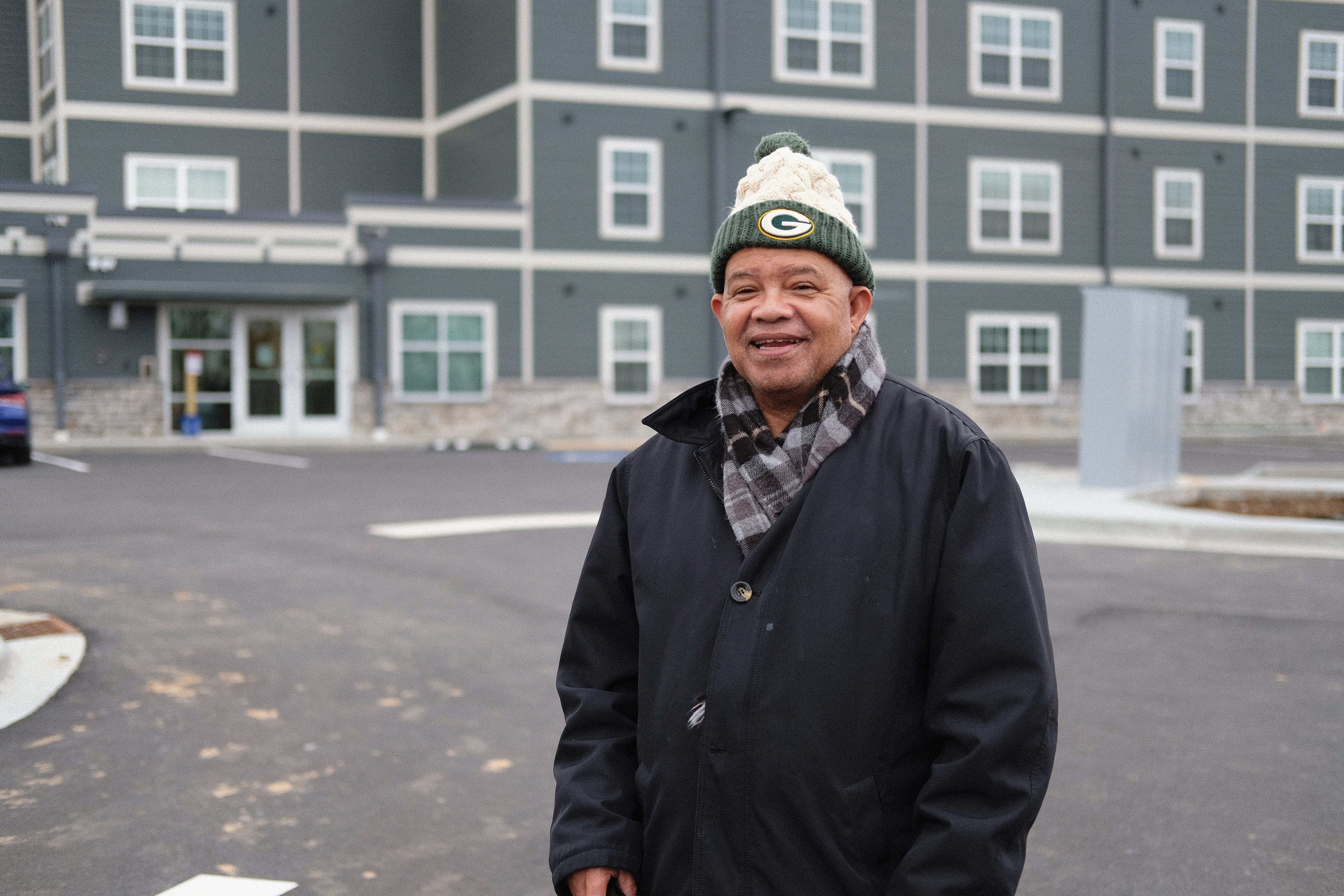 A man in a dark coat, scarf, and Green Bay Packers winter hat stands outside a gray multi-story building on a paved area.