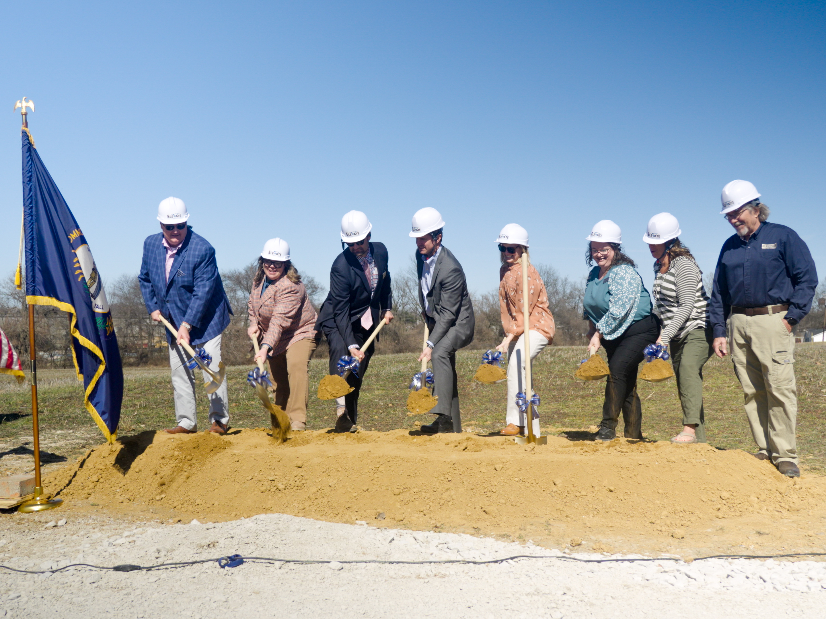 Eight people wearing hard hats participate in a groundbreaking ceremony, using shovels to dig into a mound of dirt under a clear blue sky.