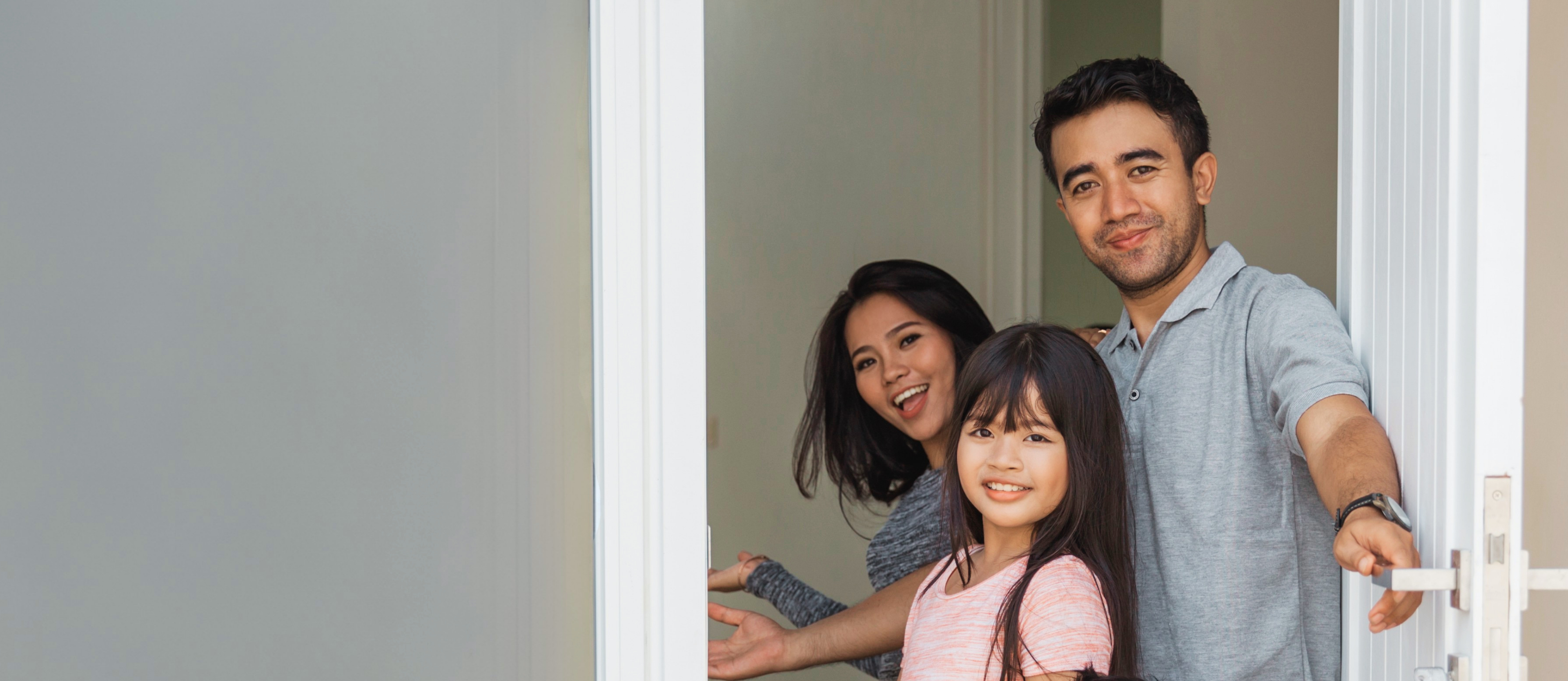 A man, woman, and young girl stand in an open doorway, smiling and gesturing for someone to enter.