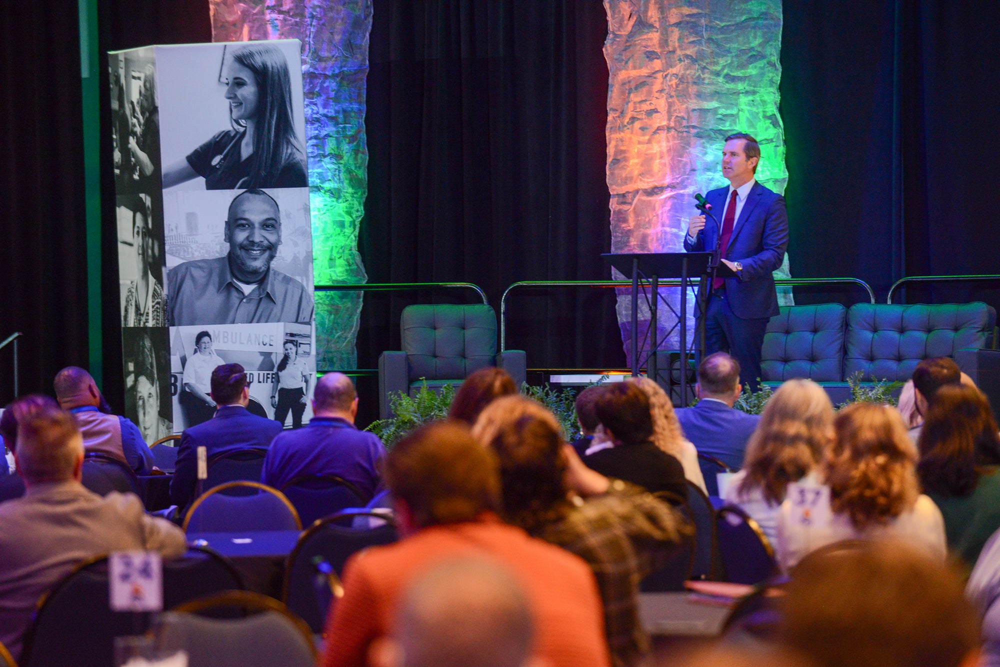 Kentucky Governor Andy Beshear addresses an audience seated at tables in a large venue with colorful lights and a backdrop featuring black-and-white portraits.