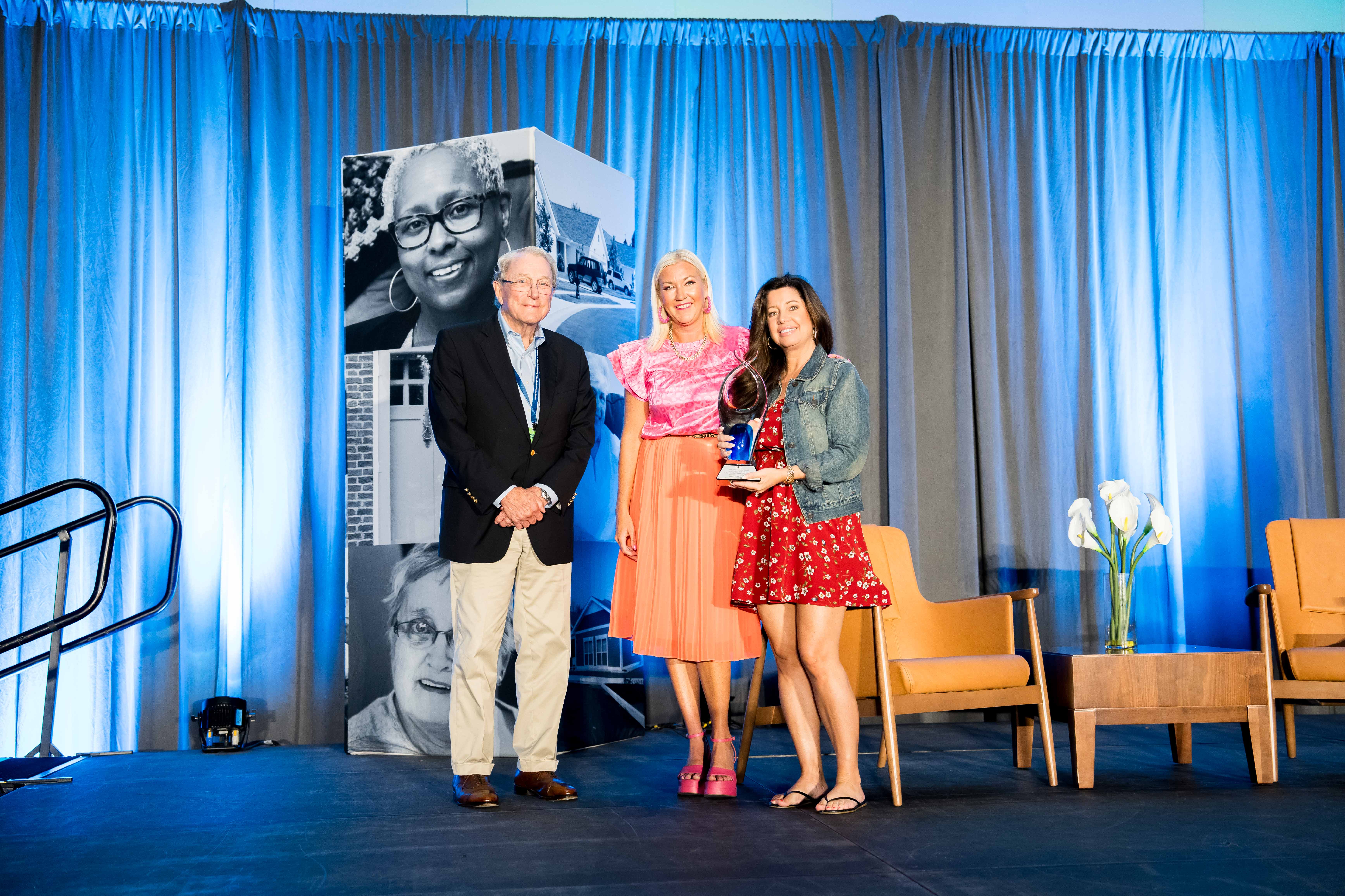 Three people stand on a stage in front of blue curtains; the woman in the center holds an award. Two empty chairs and a floral arrangement are visible on the right.