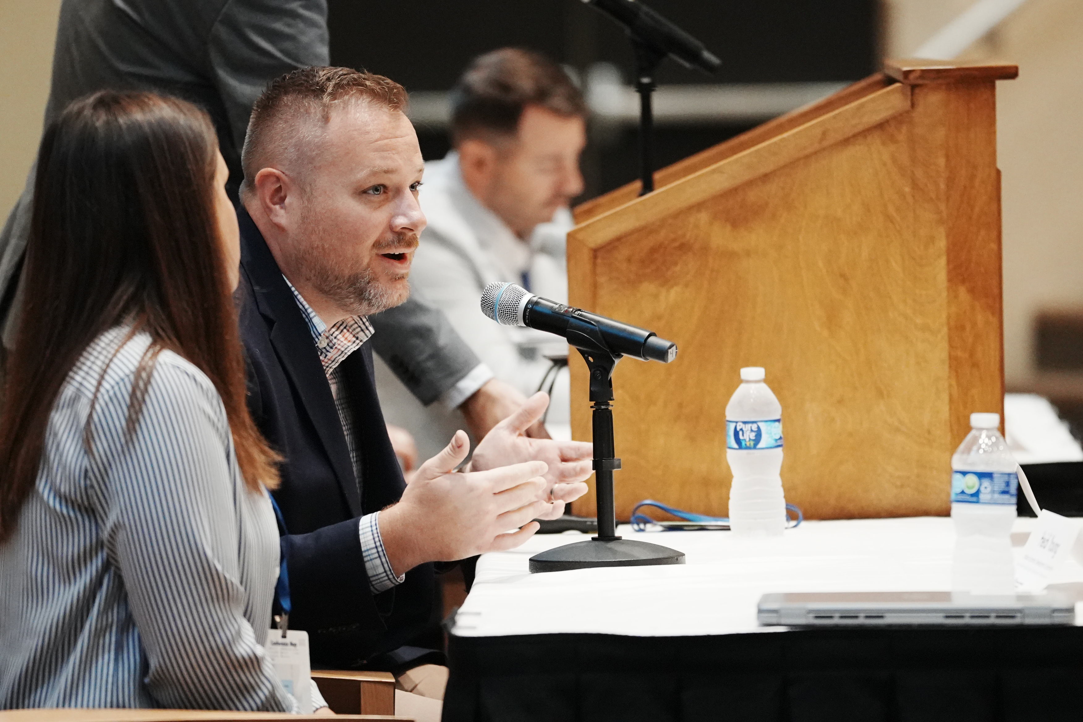 A man speaks into a microphone at a panel discussion table with water bottles, seated beside two other panelists.
