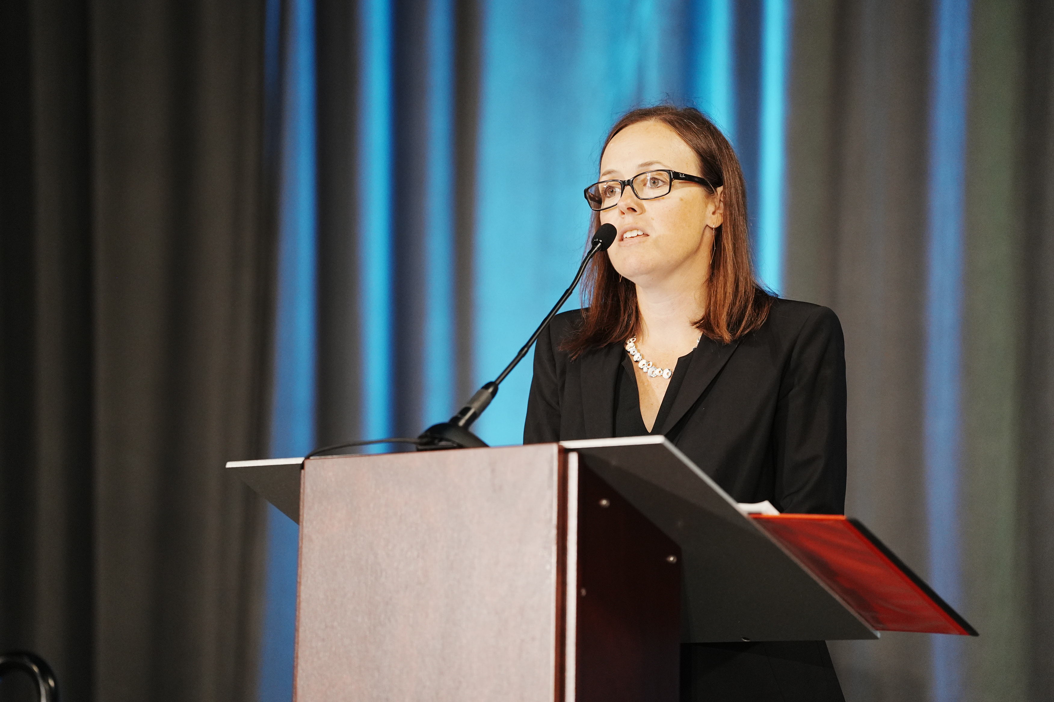 A woman wearing glasses and a black suit speaks at a podium with a microphone, standing in front of blue and gray curtains.