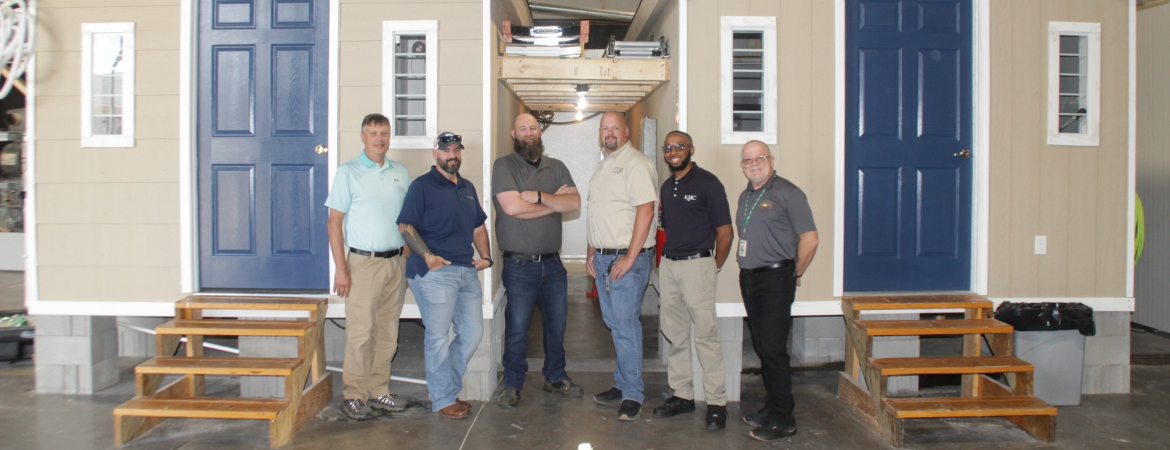 Six men stand side by side in front of two tiny houses inside a large workshop, posing for a group photo.