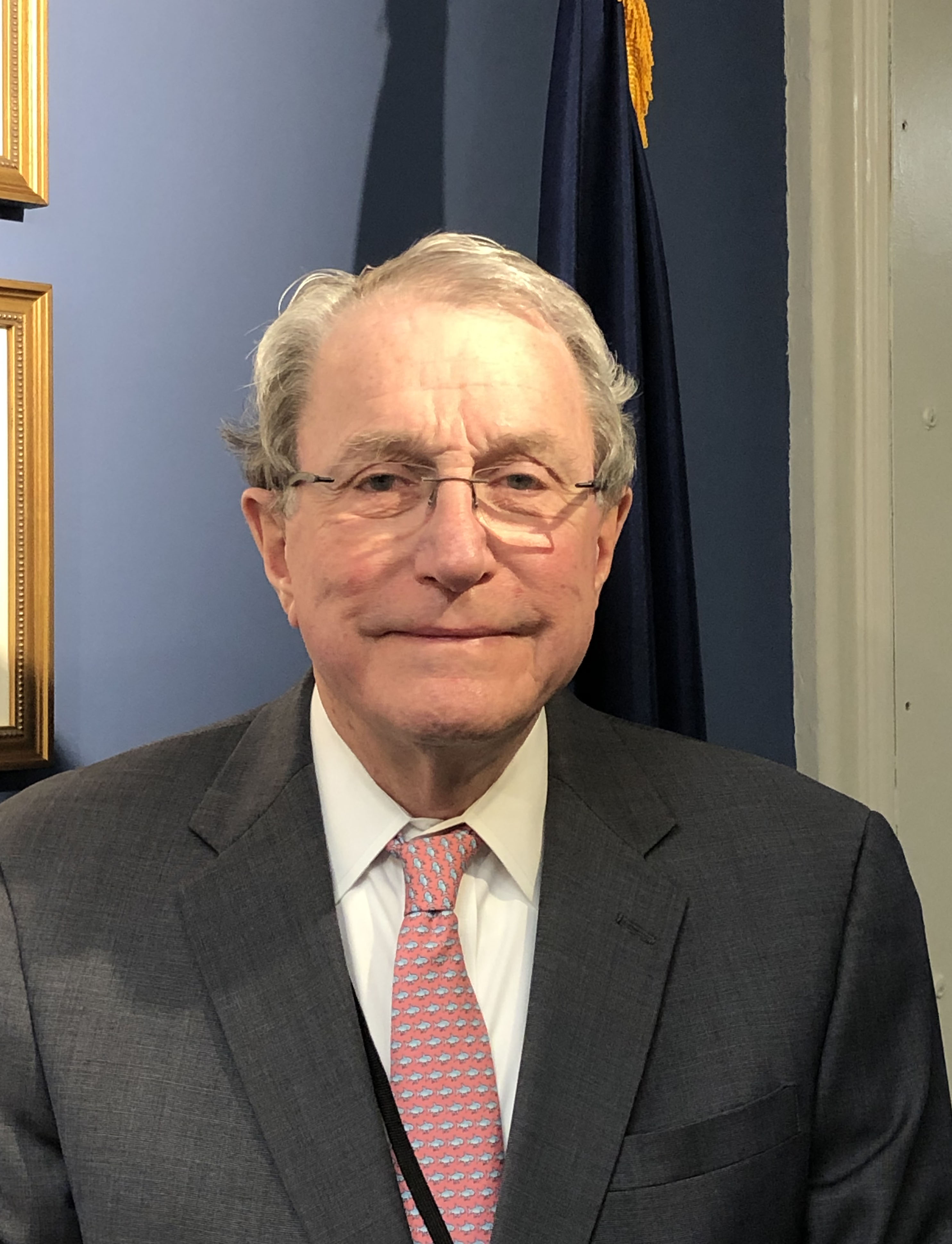 An older man in a suit and pink tie stands indoors in front of a blue wall and dark flag, facing the camera with a neutral expression.