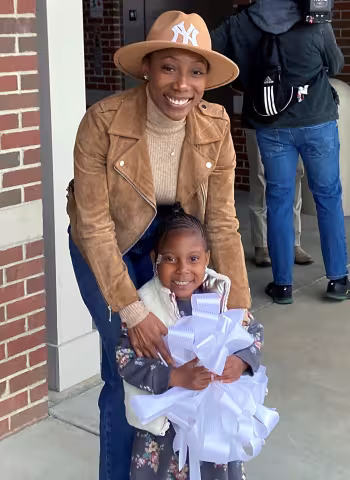 An adult and a child smile together outside a building; the child holds several white paper crafts. Both wear coats and the adult wears a tan hat.