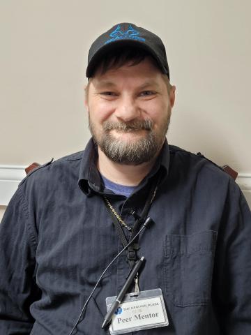 A man with a beard wearing a black button-down shirt, a baseball cap, and a Peer Mentor ID badge.