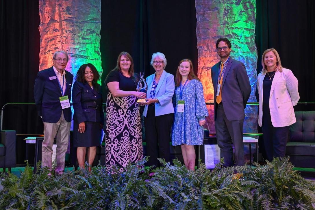 Seven people stand on a stage, some holding a trophy, with colorful lights behind them and plants in the foreground.